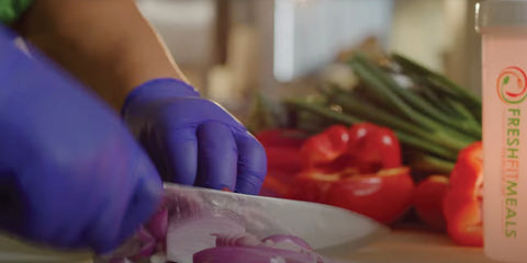 Hands in blue gloves slicing red onions, with fresh vegetables and a Fresh Fit Meals bottle in the background.